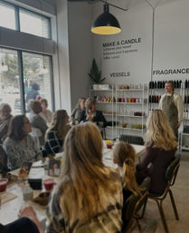 Bright candle-making workshop in a modern studio — an instructor in an apron leading a seated group around tables, with shelves of colorful vessels and fragrance bottles, large window and pendant light.