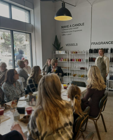 Bright candle-making workshop in a modern studio — an instructor in an apron leading a seated group around tables, with shelves of colorful vessels and fragrance bottles, large window and pendant light.