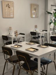 Bright minimalist studio with white tables and black padded chairs, metal trays holding paper forms and small tools at each place, framed botanical prints on the wall and a tall potted plant by the window.