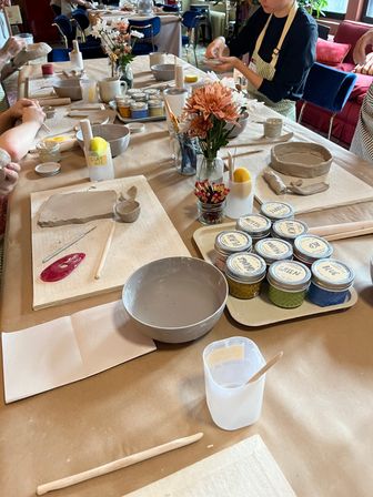 Pottery workshop table in a cozy studio—participants shaping clay with handbuilding tools, bowls of slip, labeled glaze jars, brushes and a small vase of flowers.