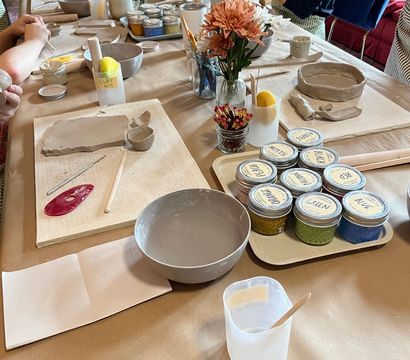 Pottery workshop table in a cozy studio—participants shaping clay with handbuilding tools, bowls of slip, labeled glaze jars, brushes and a small vase of flowers.