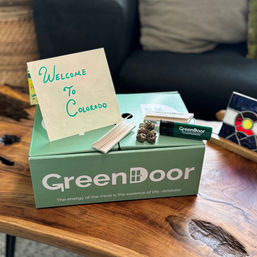 Green welcome box on a wooden coffee table with a handwritten 'Welcome to Colorado' card, cannabis buds, rolling papers and a lighter in a cozy living room setting.