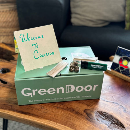 Green welcome box on a wooden coffee table with a handwritten 'Welcome to Colorado' card, cannabis buds, rolling papers and a lighter in a cozy living room setting.