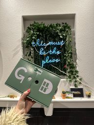 Hand holding a green box with white key and door graphics in front of a leafy wall display and blue neon sign reading "this must be the place", with small potted succulents on a white mantel