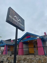Colorful cannabis dispensary storefront with a tall roadside sign, neon OPEN above a yellow door, bright pink columns and blue trim, light snow falling and a stone retaining wall in front.