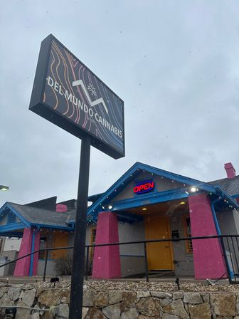 Colorful cannabis dispensary storefront with a tall roadside sign, neon OPEN above a yellow door, bright pink columns and blue trim, light snow falling and a stone retaining wall in front.