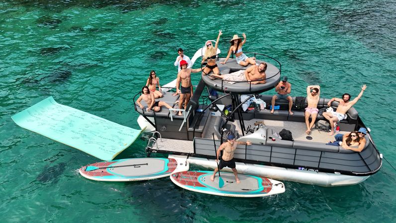 Pontoon boat party in clear turquoise water with a group of friends relaxing and waving on deck, paddleboards and a large floating mat tied alongside, sunny summer boating vibe