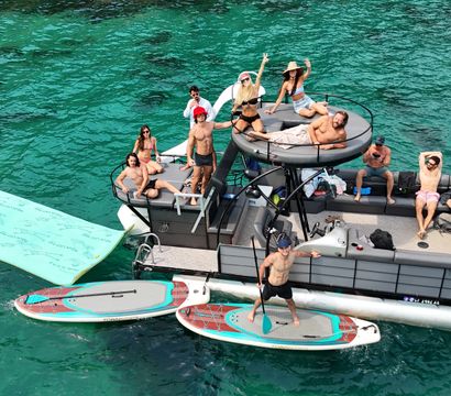 Pontoon boat party in clear turquoise water with a group of friends relaxing and waving on deck, paddleboards and a large floating mat tied alongside, sunny summer boating vibe