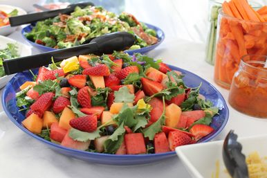 Colorful fruit and kale salad with strawberries, cantaloupe, watermelon and blueberries in a blue bowl on a buffet table with jars of carrot sticks and dressing nearby