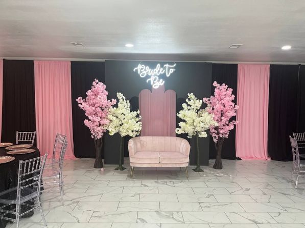 Chic bridal shower backdrop with neon “Bride to Be” sign above a blush velvet loveseat, flanked by pink and white artificial cherry blossom trees, black-and-pink drapery, clear Chiavari chairs and marble-tile event floor.