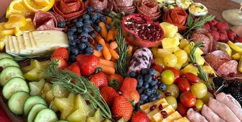 Colorful party-ready charcuterie and fruit platter with strawberries, blueberries, grapes, pomegranate, pineapple, cucumber and starfruit slices, cherry tomatoes, rolled salami, sliced cheeses, baby carrots, deviled eggs and rosemary sprigs.