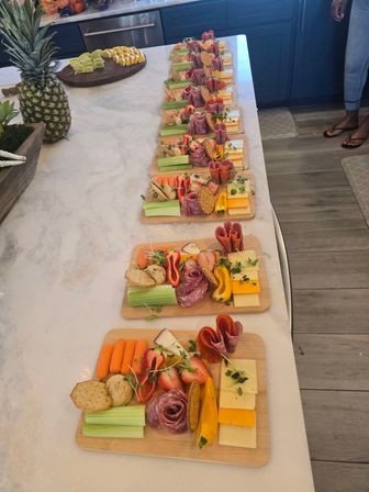 Colorful wooden charcuterie boards lined up on a white kitchen island, each with rolled salami and pepperoni, stacked cheeses, crackers, celery sticks, baby carrots, strawberries, yellow bell pepper and microgreens — ready-to-serve party appetizers.