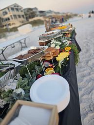 Beachside buffet on a sandy shore with foil trays of grilled burger patties and buns, platters of sliced pineapple, oranges, dragon fruit and strawberries, decorative greenery and stacked white plates with coastal houses blurred in the background.
