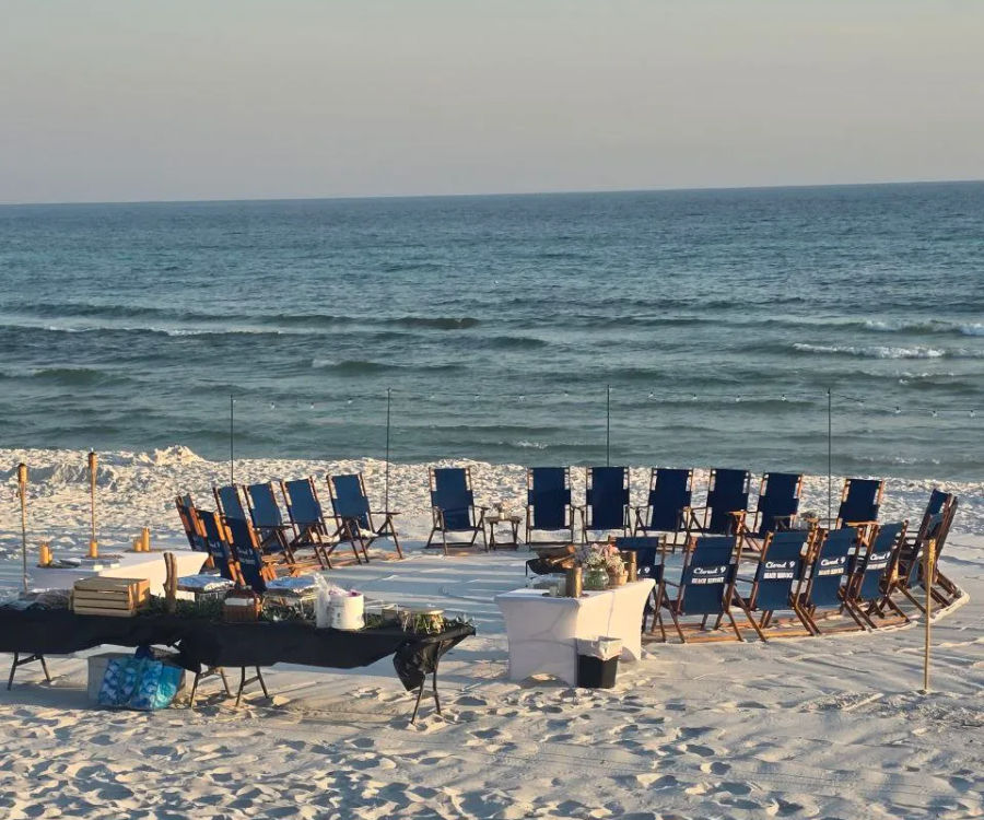 Blue folding chairs arranged in a circle on white sandy beach facing calm ocean waves, with tables, tiki torches and event supplies set up for a seaside gathering.