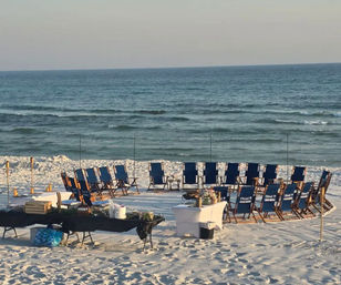 Blue folding chairs arranged in a circle on white sandy beach facing calm ocean waves, with tables, tiki torches and event supplies set up for a seaside gathering.