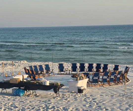 Blue folding chairs arranged in a circle on white sandy beach facing calm ocean waves, with tables, tiki torches and event supplies set up for a seaside gathering.