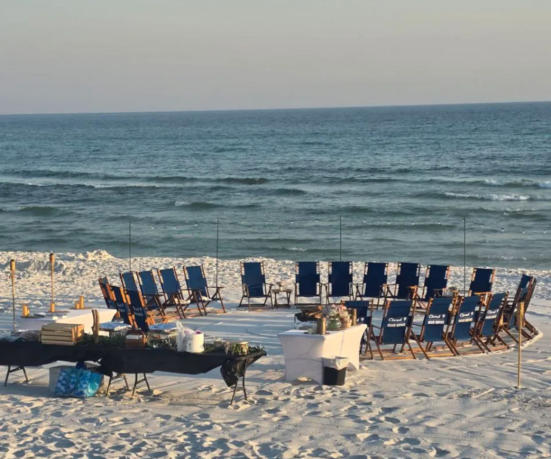 Blue folding chairs arranged in a circle on white sandy beach facing calm ocean waves, with tables, tiki torches and event supplies set up for a seaside gathering.