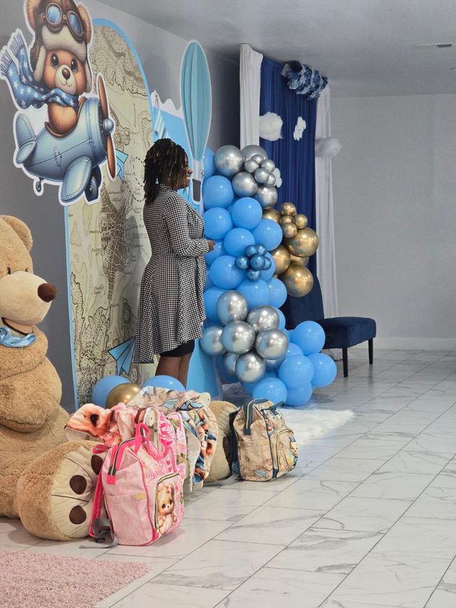 Indoor airplane-and-teddy themed baby shower display with blue-and-gold balloon garland, vintage map backdrop, large plush bear, colorful kids' backpacks on marble floor and a person standing beside the setup