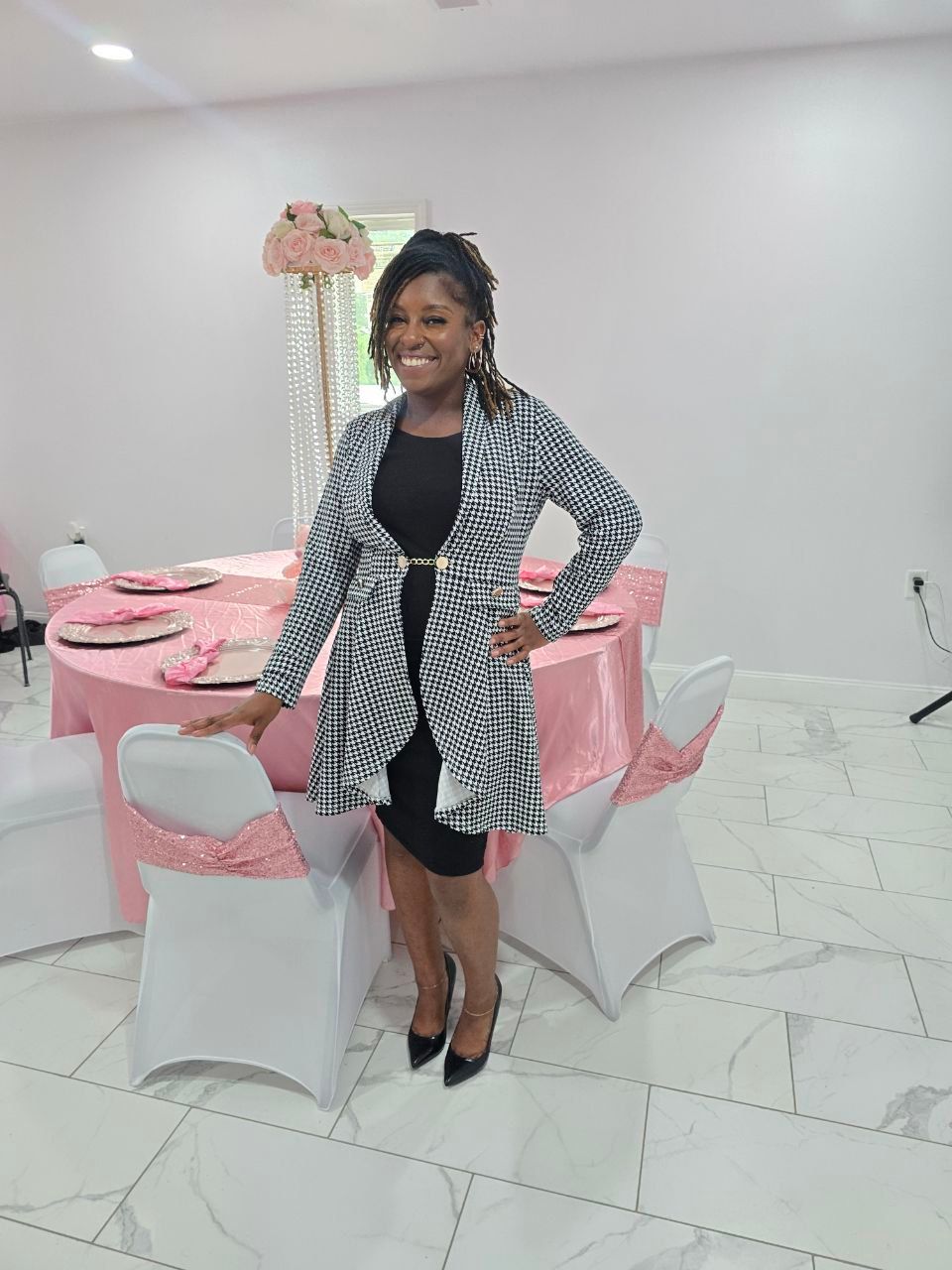 Smiling woman in houndstooth blazer and black dress posing by a pink-and-white banquet table with rose centerpiece in an indoor event venue