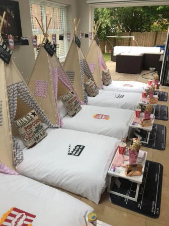 Backyard-facing living room set up for a kids' movie-night sleepover with four teepee beds lined up, white bedding, popcorn-themed pillows, clapperboard mats, string lights and bedside trays holding popcorn and drinks.