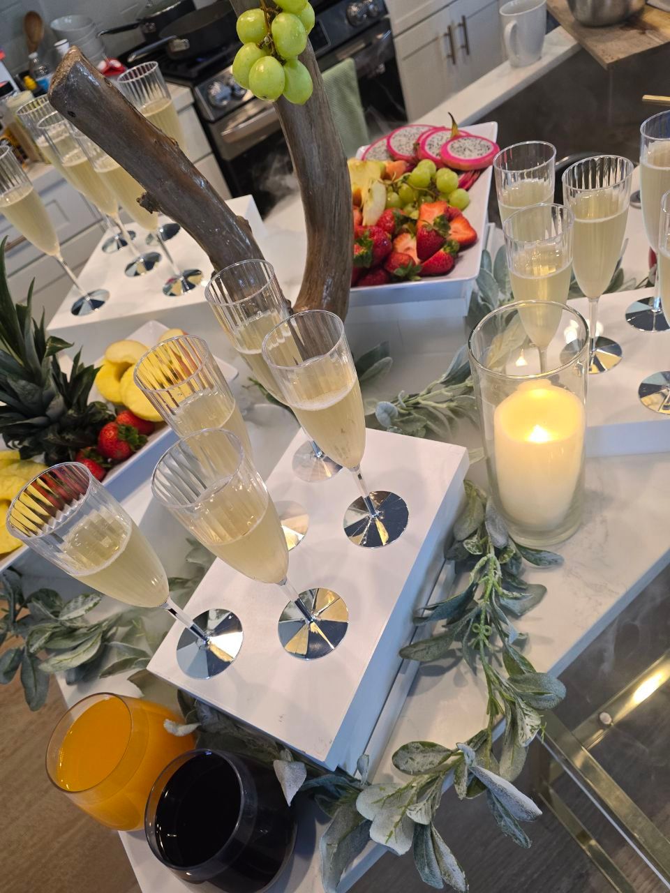 Cheerful brunch spread on a modern kitchen island: champagne flutes of sparkling wine, a fruit platter with strawberries, grapes, dragon fruit and pineapple, orange juice, coffee and a lit candle with greenery accents.