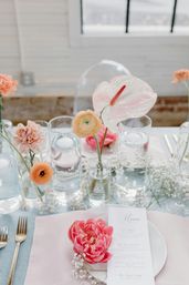 Sunlit indoor wedding tablescape with a bright pink peony on a menu card, glass vases holding pale anthurium and ranunculus, baby's breath, and gold flatware on pastel linens.