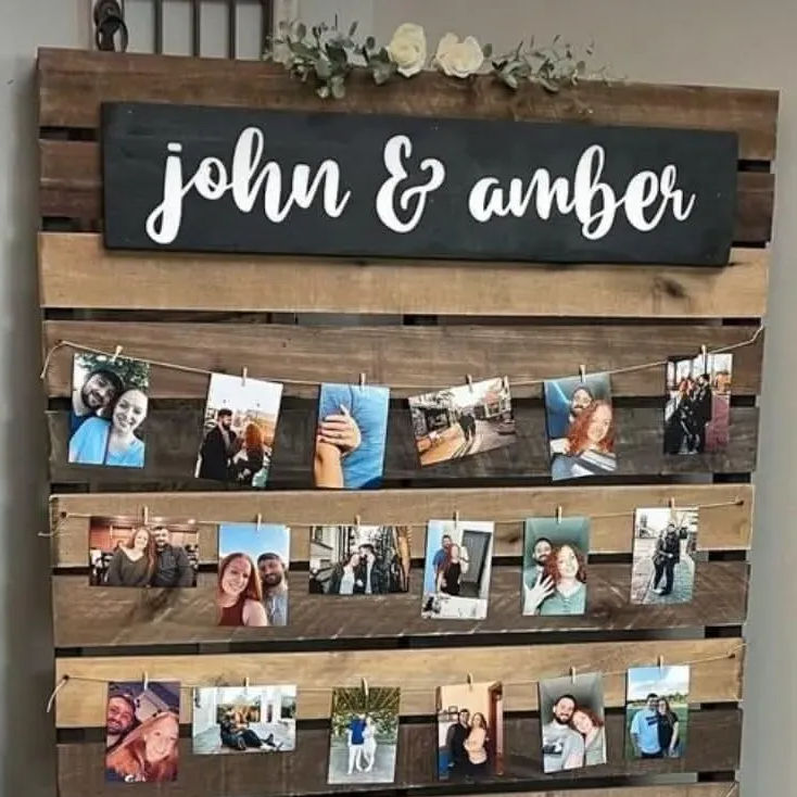 Rustic wooden pallet photo display topped with a black sign reading 'john & amber' and cream flowers, with three rows of twine and clothespin‑hung couple photos — DIY wedding/engagement memory board.
