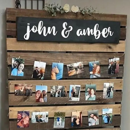 Rustic wooden pallet photo display topped with a black sign reading 'john & amber' and cream flowers, with three rows of twine and clothespin‑hung couple photos — DIY wedding/engagement memory board.