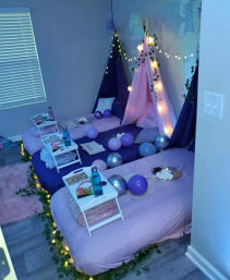 Cozy indoor slumber party setup with pink and purple floor beds and teepee tents draped in fairy lights, metallic balloons, ivy garlands and individual snack trays for a fun sleepover.