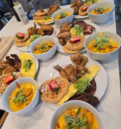 Vibrant brunch spread on a white countertop: chicken and waffles with strawberry, fried chicken, omelettes, crispy bacon and bowls of creamy shrimp curry topped with microgreens.