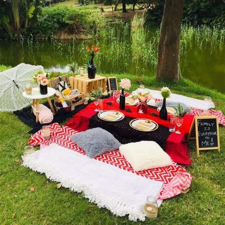 Cozy lakeside picnic spread on a grassy park bank with patterned blankets, fluffy cushions, tableware and wine bottles, floral arrangements, and a small chalkboard reading Family is everything to me.