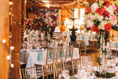 Elegant rustic barn wedding reception with lace-covered tables and teal runners, gold Chiavari chairs, tall pink and white floral centerpieces, sparkling string lights and glassware set for guests