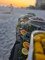 Beachside buffet at sunset on a sandy shore — table with foil trays, corn on the cob, sliced oranges and limes, and greenery garnish with a blurred seaside background.
