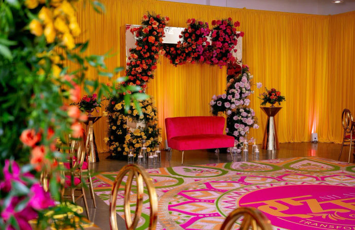 Vibrant indoor event stage with a pink velvet loveseat beneath a lush floral arch of red, pink and yellow roses, golden accent chairs, candle votives and a bright yellow draped backdrop over a colorful circular patterned floor decal.