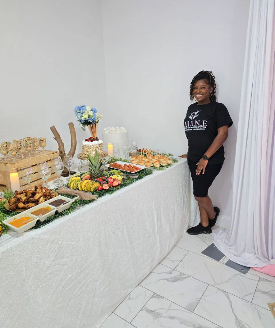Smiling staff member beside an indoor catered buffet with sliders, chicken wings, fresh fruit display, desserts, flowers and candles on a white draped table.