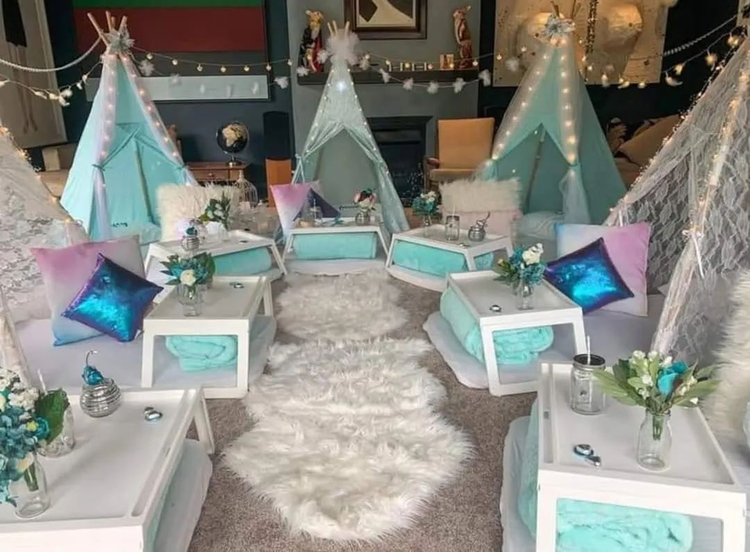 Indoor kids' teepee slumber party setup with mint-turquoise mini tents, fairy lights, fluffy white rugs, low white tables, metallic pastel pillows and small floral centerpieces.