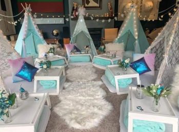 Indoor kids' teepee slumber party setup with mint-turquoise mini tents, fairy lights, fluffy white rugs, low white tables, metallic pastel pillows and small floral centerpieces.