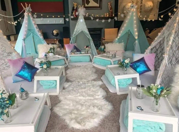 Indoor kids' teepee slumber party setup with mint-turquoise mini tents, fairy lights, fluffy white rugs, low white tables, metallic pastel pillows and small floral centerpieces.