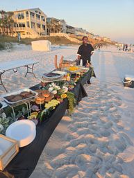 Sunset beachfront buffet on white-sand shore with a long table of burgers, fresh fruit and chafing trays, server standing by, coastal vacation homes on the dunes in the background.