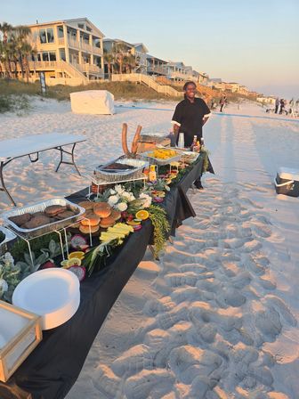 Sunset beachfront buffet on white-sand shore with a long table of burgers, fresh fruit and chafing trays, server standing by, coastal vacation homes on the dunes in the background.