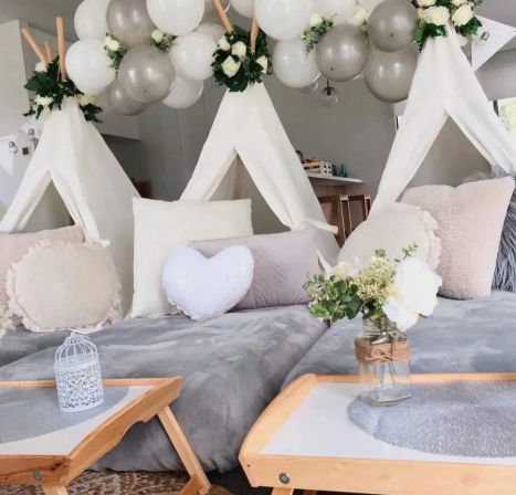Cozy indoor home sleepover setup with three white teepee tents, gray plush bedding, heart and round throw pillows, silver-and-white balloon garland, wooden trays and mason jar flower centerpiece