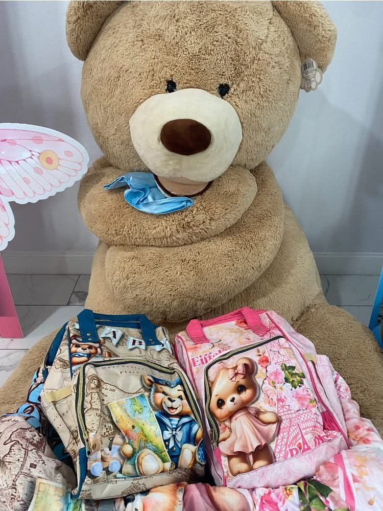 Large tan teddy bear cuddling two kids' backpacks: a blue map-themed bear-print backpack and a pink floral teddy-print backpack on a display