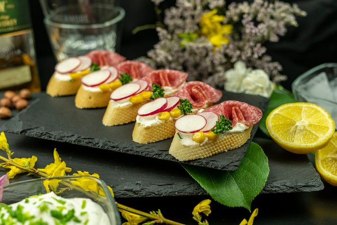 Row of gourmet crostini canapés on a black slate board: baguette slices topped with cream cheese, sliced radishes, corn kernels, parsley and folded salami, styled with lemon halves and spring flowers