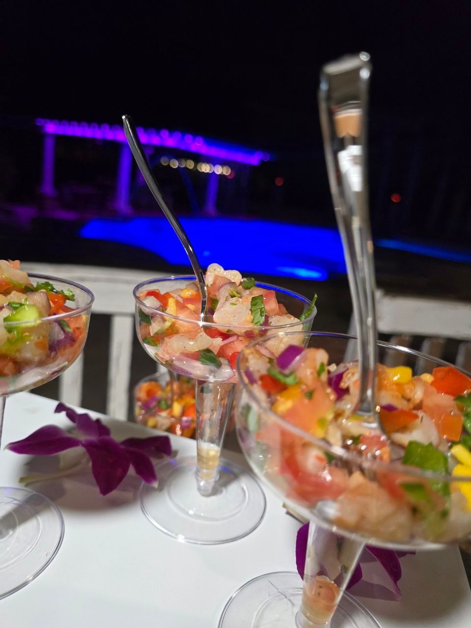 Colorful shrimp ceviche in martini glasses with diced tomato, red onion, cilantro and mango, orchid garnish on a table poolside at night with blue-purple LED-lit pool in the background.