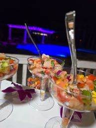 Colorful shrimp ceviche in martini glasses with diced tomato, red onion, cilantro and mango, orchid garnish on a table poolside at night with blue-purple LED-lit pool in the background.