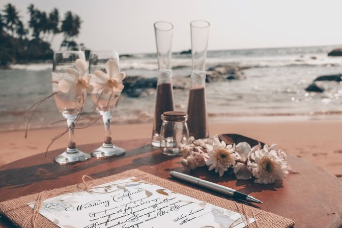 Wedding certificate on a wooden table at a tropical palm-lined beach, with two flower-adorned champagne flutes, a pen, glass vases and a floral lei against the ocean waves