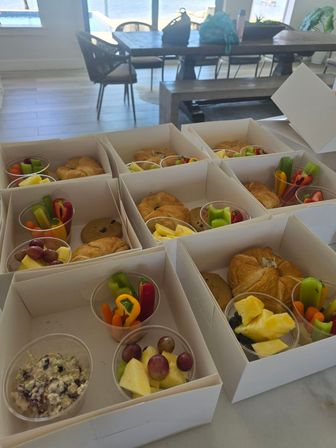 Neatly arranged catered breakfast boxes with croissants, cookies, fruit cups and veggie cups on a kitchen island in a bright waterfront dining room