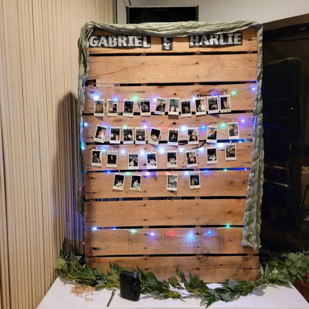 Rustic wooden pallet photo display at an indoor wedding reception, Polaroid-style photos clipped to string lights with multicolored fairy lights and greenery on the table.