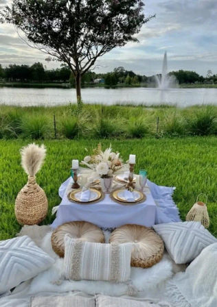Boho lakeside picnic: low white-draped table set with gold chargers, dried floral centerpiece, candles and glassware, surrounded by cream cushions, wicker lanterns and pampas grass on a grassy shore with a lake fountain and tree.