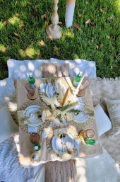 Sun-dappled backyard picnic tablescape on green lawn with low wooden table, neutral boho place settings, woven placemats, white plates and gold cutlery, pampas grass centerpiece, green glass bottles, pink goblets and cushions on blankets